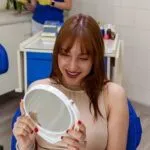 Smiling young woman looking into a handheld mirror in a modern dental clinic, satisfied with the results of her dental procedure or smile makeover